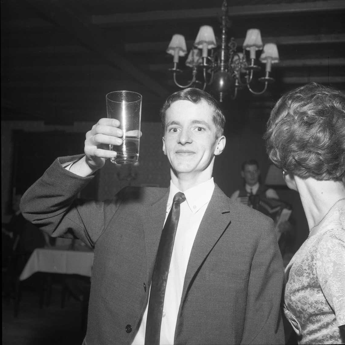 En ung och glad man i kostym och slips lyfter ett glas som en skål mot fotografen. En kvinna till höger, delvis ur bild, är vänd med blicken åt andra hållet. Det är ett fotografi i samband med årsmöte för Gränna AIS.