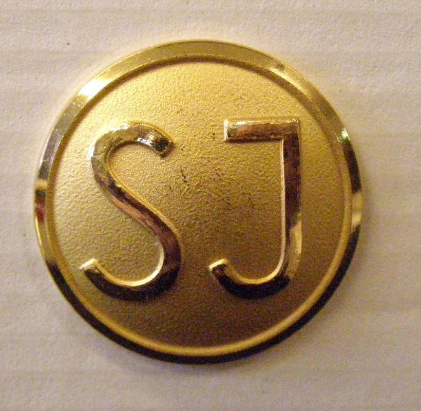 Guldfärgad rockknapp med SJ:s logotyp i form av enkla initialer.