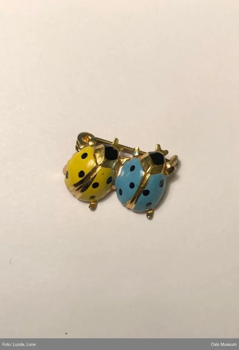 Nål i metall i form av to "marihøner" med emaljedekor i blått og gult.
