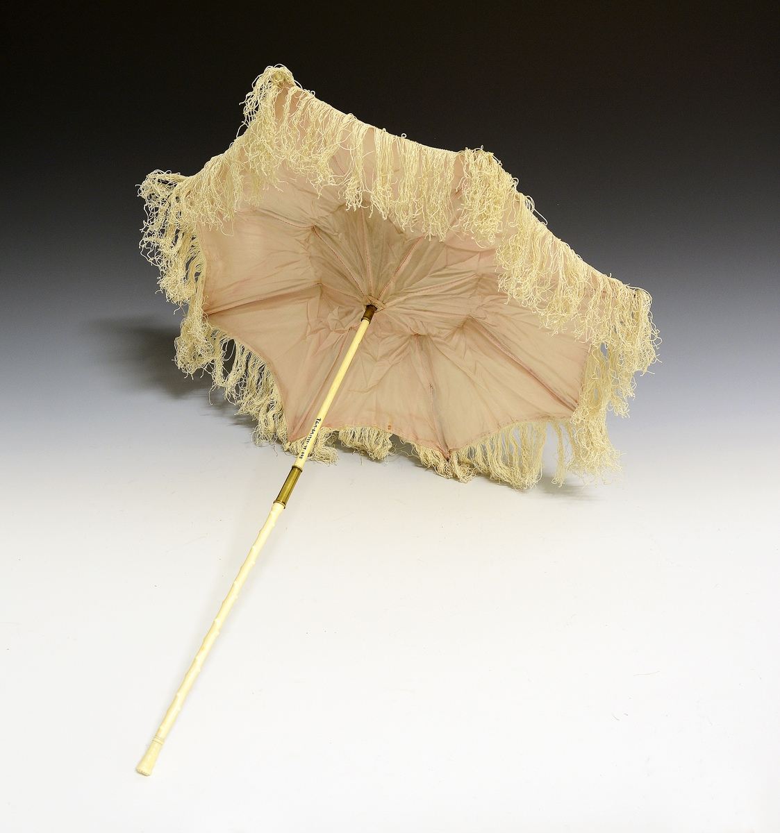 Knekkparasoll. Fra protokollen: "Knekk-parasol" av kvit silke, med bleikraudt fôr og tafs ikringum. Stongi, som er av elfenbein hev massingholk til aa smøygje yvi leden, og beint, knuprut skaft paa lag 48 cm. langt. Tre-spilur.