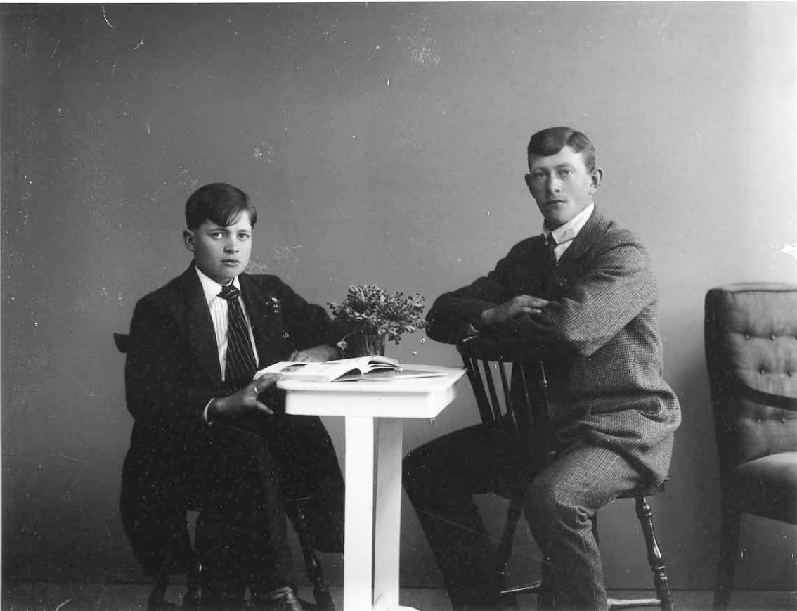 Gruppbild med två unga män sittande vid ett bord, med en uppslagen bok. Sven Blomkvist till vänster med randig slips. Algot Lätt till höger sitter bakvänd på en stol och vilar armarna på dess ryggstöd.