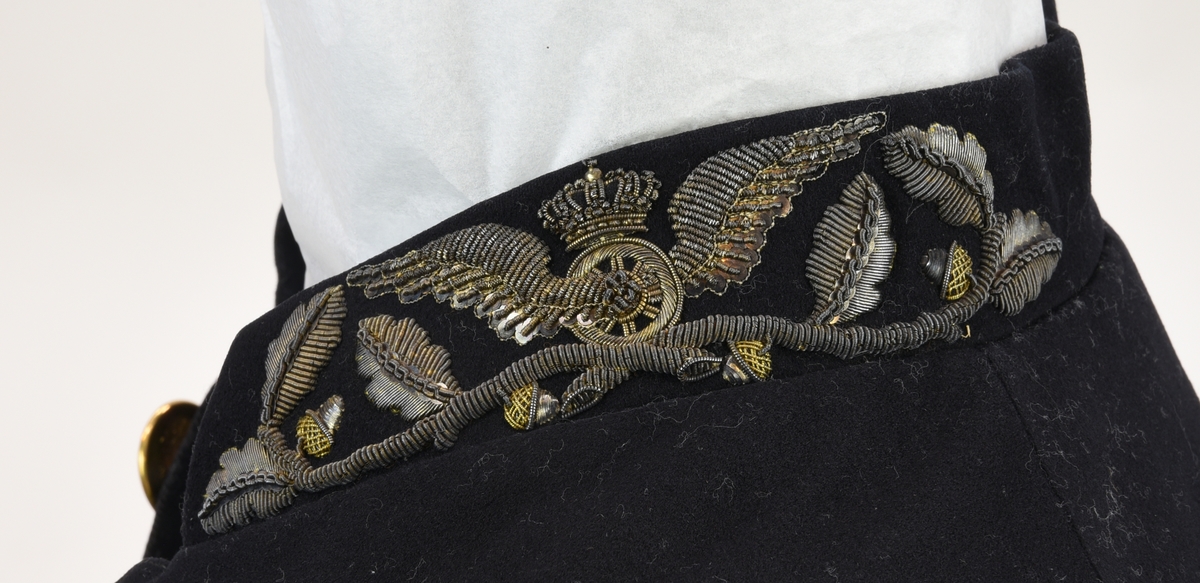 Enkelknäppt paradfrack av mörkblått kläde med liten ståndkrage med metallbroderier i form av vinghjul och enklare eklövsrankor. Fracken har svängd mitt fram-linje och sju guldfärgade knappar.