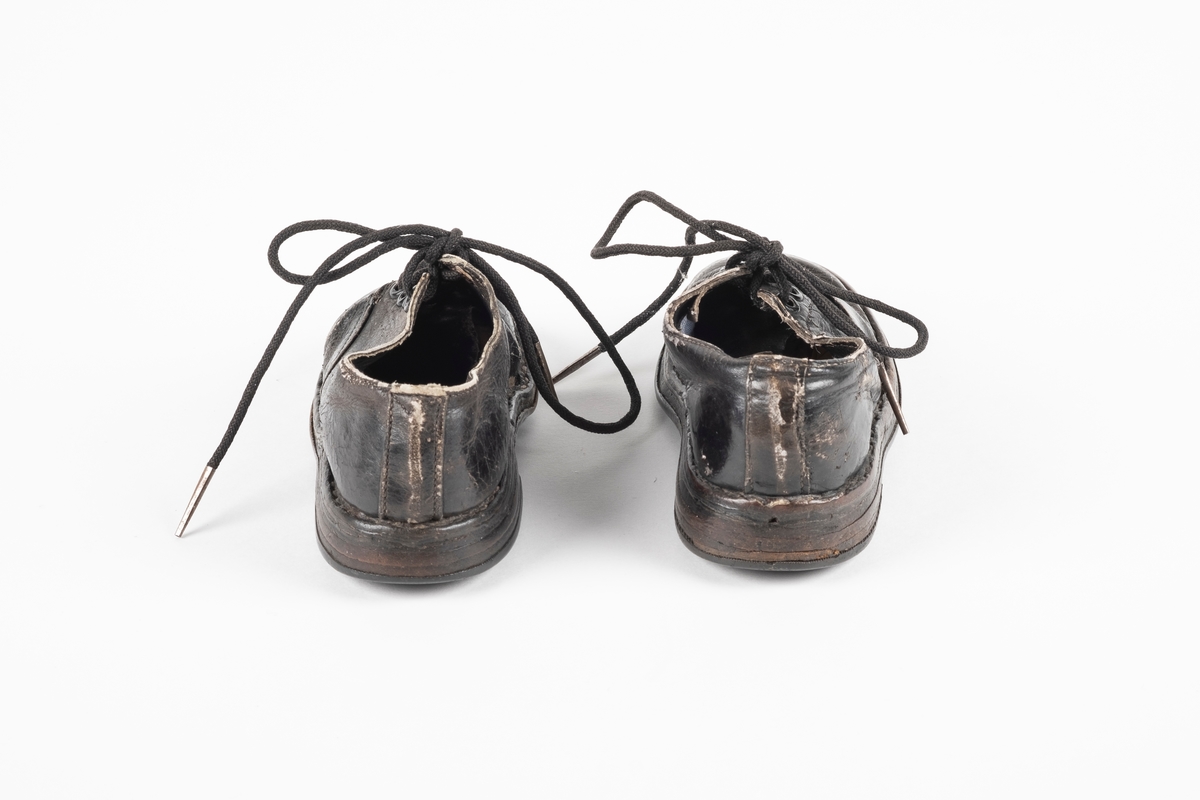 Ett par randsydd barnesko av lær. Skoene har snøring med runde lisser. Maljene er av metall. Sålen er av lær og er forsterket med spiker på hælen og ved tuppen på undersiden.