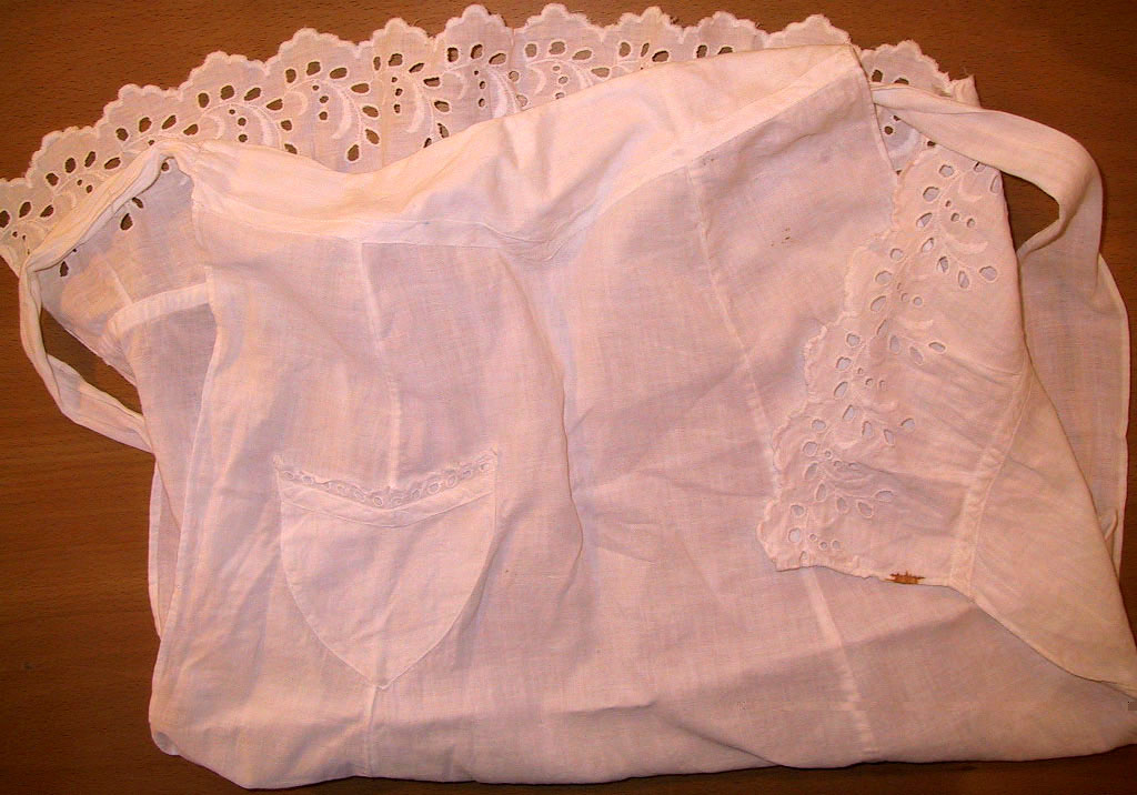 Hvitt bomullsforkle med knytebånd og lomme. Pyntet med blondekant nederst og på lommen.
