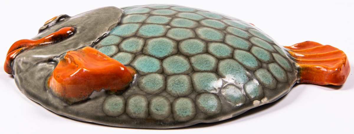 Väggfigurin i lergods i form av en fisk. Modell H.12 (angivet på baksidan) eller HO12 (enligt förteckning), glasyr 508 i orange, blekgrön och ljusbrun. Glasyren 508 användes enligt Birgitta Lundblads bok 1934-48.
