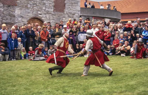 To riddere i rustning og med røde overkapper utkjemper blankvåpenkamp foran Storhamarlåven. Mellom ridderne og låven er det en stor menneskemengde som ser på kampen.. Foto/Photo