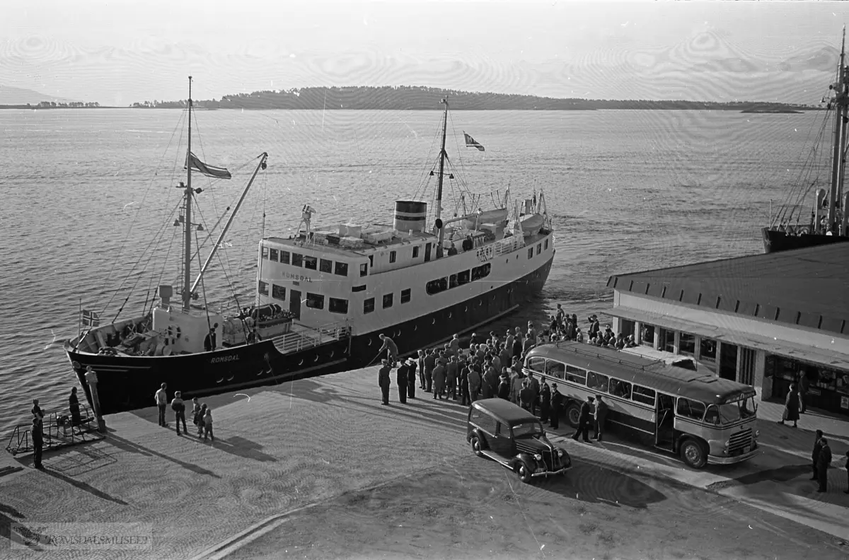 "September 1958"."Oksviks gravferd".Fra begravelsen til Olav Oksvik f.07.05.1887 d.16.09.1958..Båten som legger til kai er M/S Romsdal, som gikk i rute Åndalsnes-Molde-Ålesund i tidsrommet 1950-1962. Den ble solgt i 1962..Bussen på bildet er sannsynligvis T-8685 Scania-Vabis, 1953-modell, med karosseri fra Bussbygg, Hovdenakken. Bussen tilhørte Aarø Automobilselskap (Auto) og den var lakkert i gult og mørk blå. Aarø Auto hadde ruter Molde-Kristiansund over Gjemnes, og ruta startet og avsluttet ved Storkaia i Molde, der det var korrespondanse med båter til Åndalsnes og Ålesund. .Begravelsesbilen er en Ford V8 1936 med registreringsnummer T-2167.