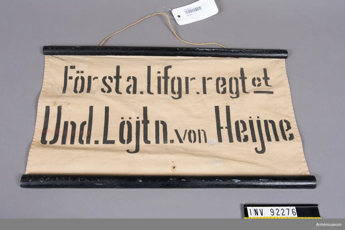 Tygskylt fäst mot två svartmålade träribbor. Tryckt text: "Första Lifgr.regt:et Und. Löjtn. von Heijne".