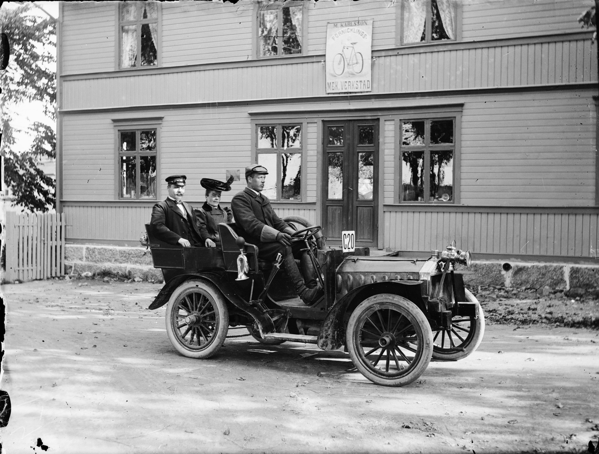 Mattias Karlsson i sin bil med en kvinna och chaufför Gustav Lundström utanför "M. Karlsson förnicklings Mekaniska Verkstad", Östhammar, Uppland 1915