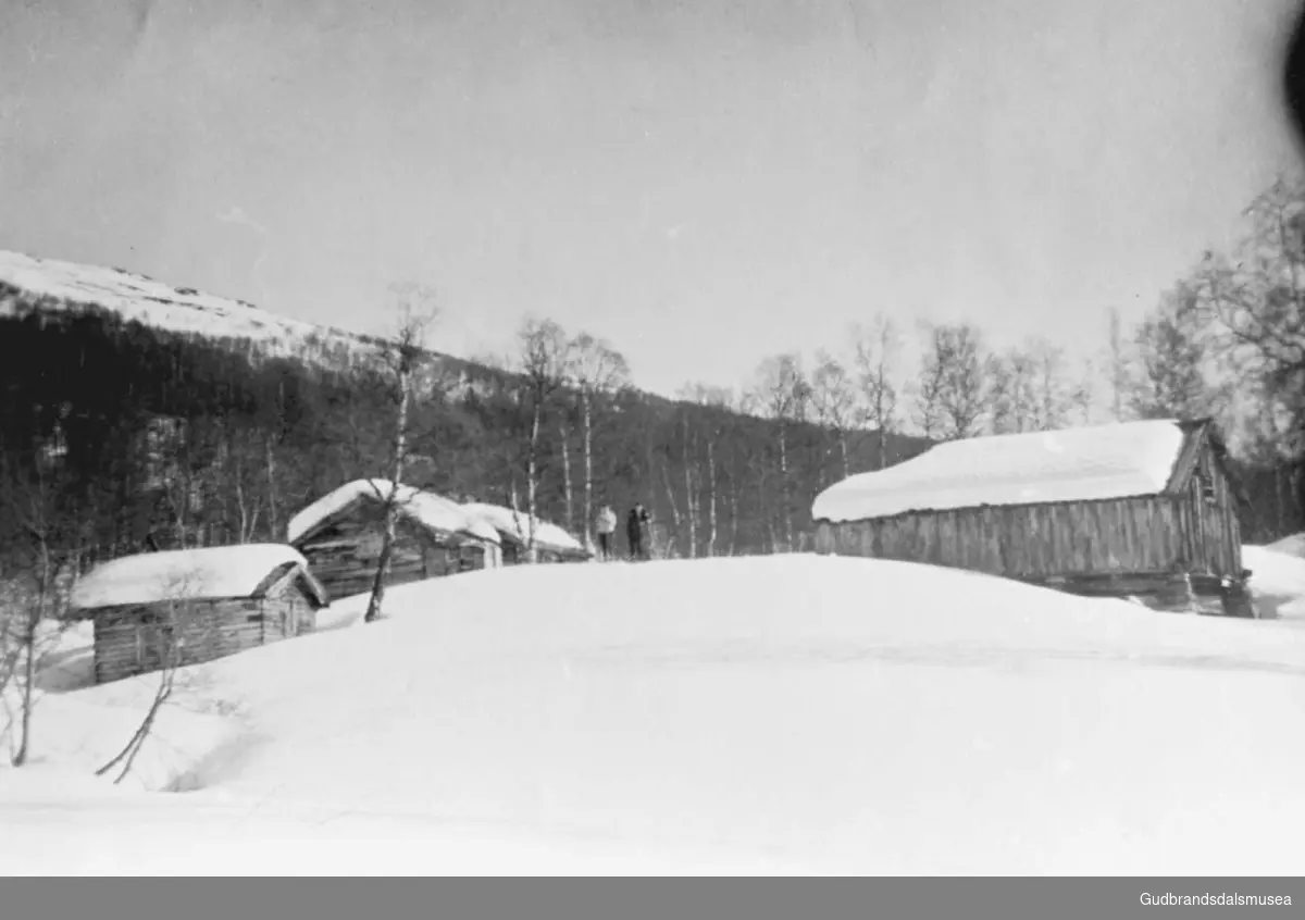 Mindre bygninger, fra venstre kokkhus Uppistugu, selet til Uppistugu, selet til Nistugu, fjøset til Uppistugu, to personer står mellom bygningene, de går på ski?, det er vinter og snø. 