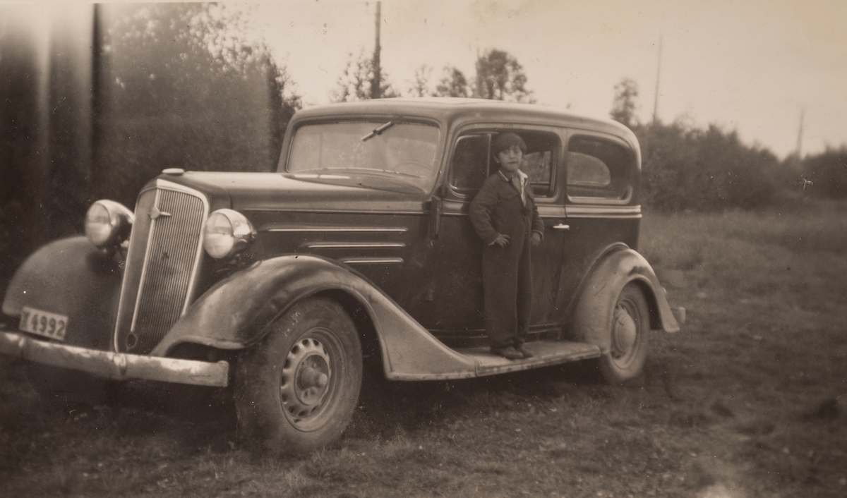 En pojke står på tröskeln till en bil, lutad mot förarsidans dörr. Han är klädd i svart overall och håller händerna i sidorna. Pojken och bilen finns i Kungsgården 1946.