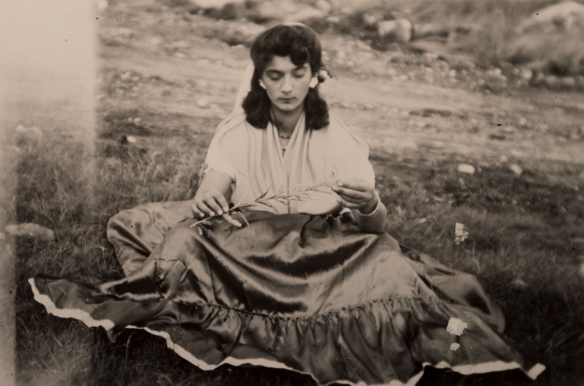 Romsk kvinna sitter på marken en sommardag i Hofors, 30 juli 1950. Hon tittar på en växt som hon håller i händerna.