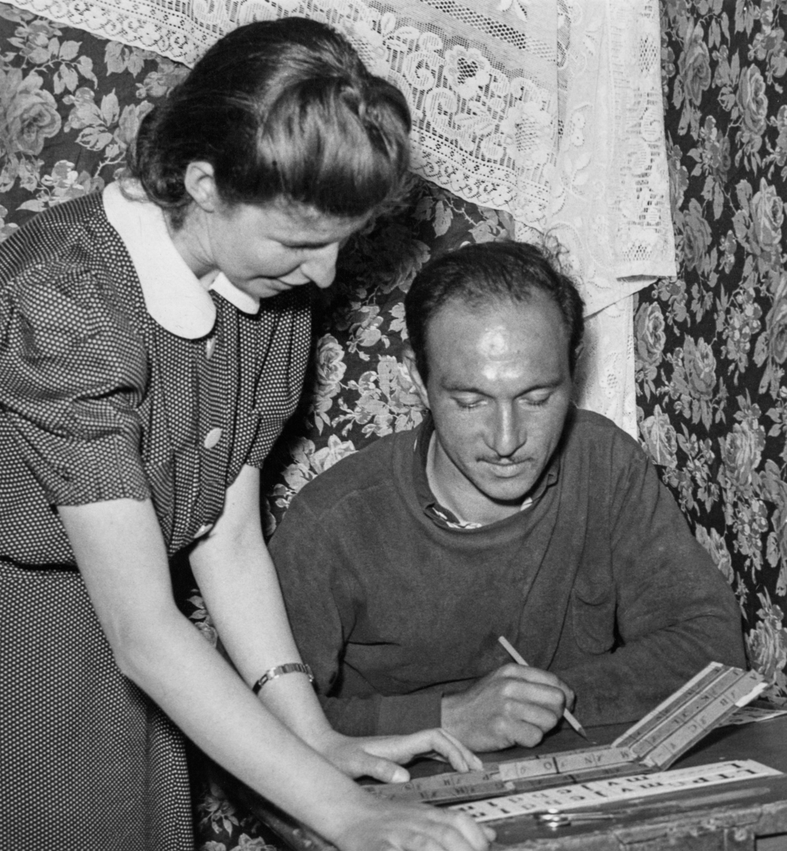 Skolfröken hjälper en romsk elev att läsa sig läsa och skriva under sommarskola 1943. Mannen sitter i bänken och håller i en penna.