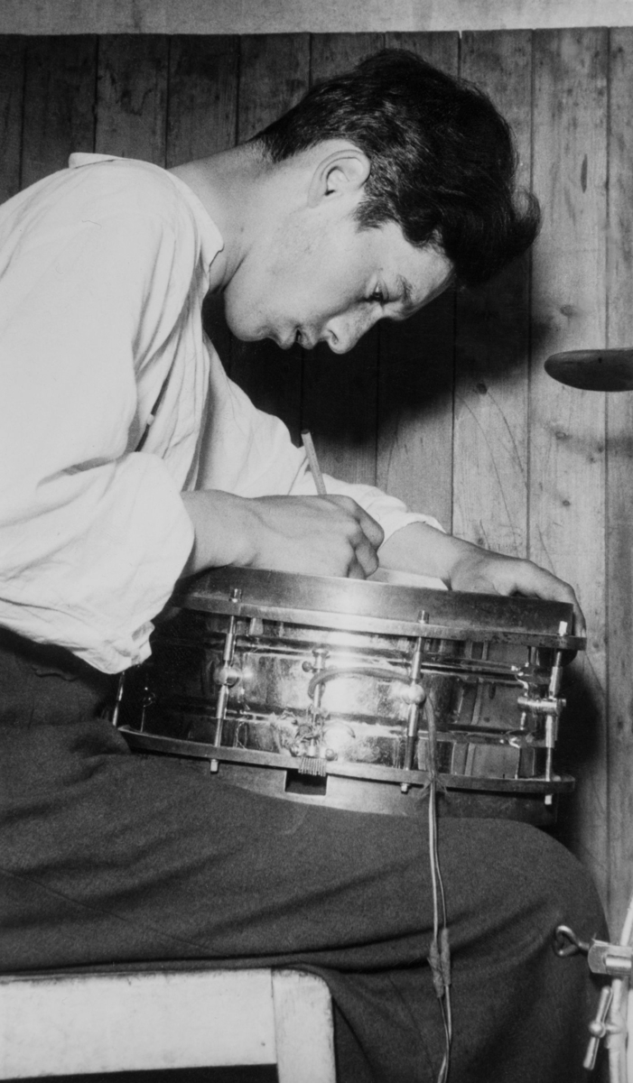 En ung man skriver något med sin virveltrumma som underlag. Ett cymbalstativ skymtar i bildens ytterkant.