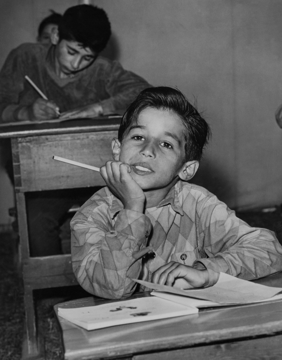 En pojke sitter i sin skolbänk med hakan stödd i handen. Bakom honom skriver en annan pojke flitigt i sin bänk.
