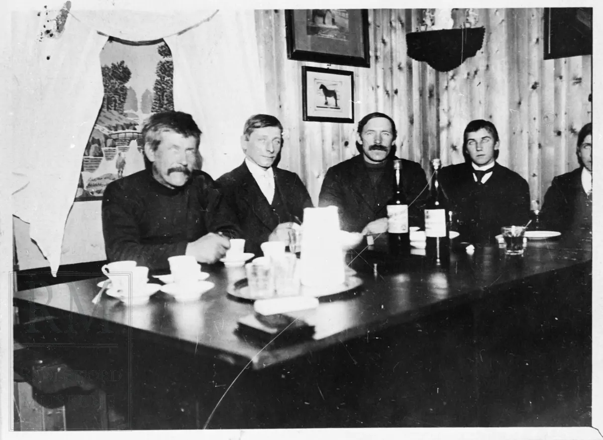 5 menn sitter rundt et bord. Kopper, glass og flasker på bordet.