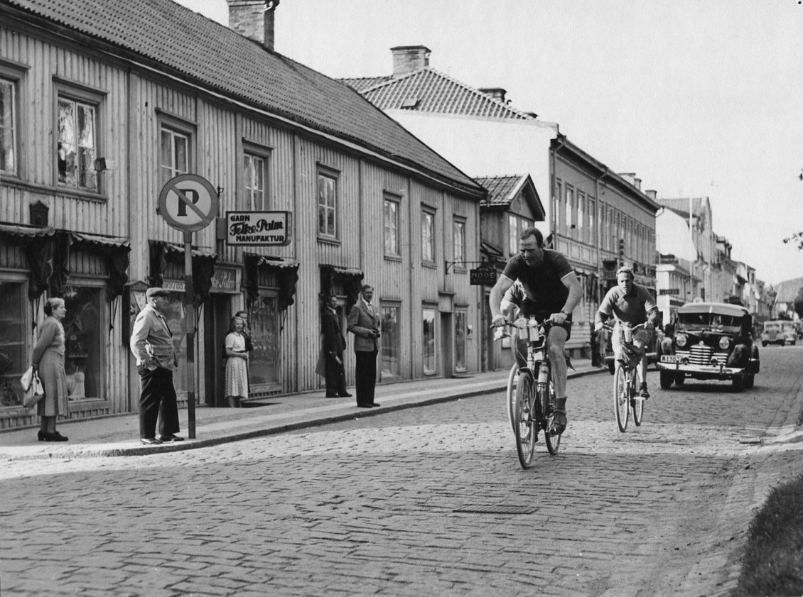 Boxaren Olle Tandberg med två cyklister efter sig på Brahegatan i Gränna, följda av bilar, i samband med Sverigeloppet. Enstaka publik på trottoraren utanför Folke Palms Garn- och Manufakturaffär.