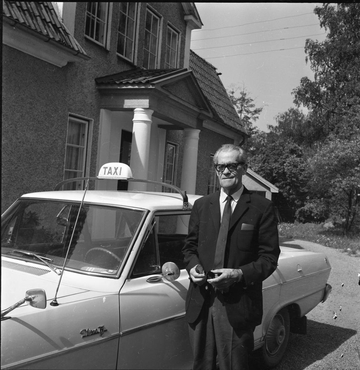Droskägaren Oskar Blomqvist står vid sin taxibil - en Chevrolet Nova - utanför en villa. Han håller solglasögon i händerna.