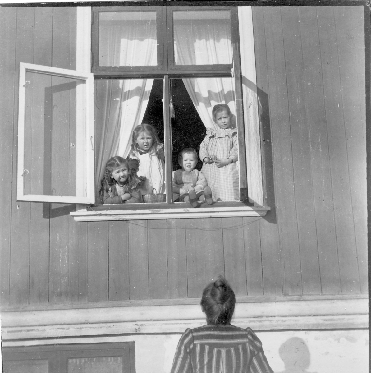 En kvinne står og ser opp på fire barn som står i et åpent vindu.