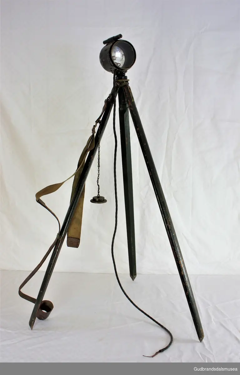 Signallampe med trefot stativ.Komplett med bærereim, med kabel som mangler stikker.
Brukt på Otta i Gudbrandsdalen under kampene der.
Lampa er merket 1918, og stativet London 1917.