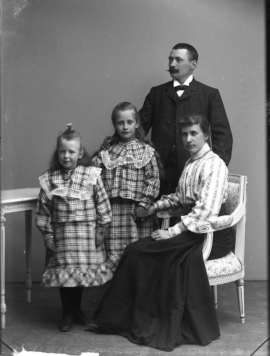 Gruppbild med familjen Franzén i Gränna. Två flickor i storrutiga klänningar med volang står intill deras mamma som sitter i en stol av gustaviansk stil. Flickan närmast mamman (den äldsta) håller hennes hand. Mannen står bakom dem. Hans högra hand vilar på den äldre flickans högra axel. Han tittar mot vänster, de andra tittar på fotografen.
Från vänster: Linnea, Gerda, Olga samt Gustaf.