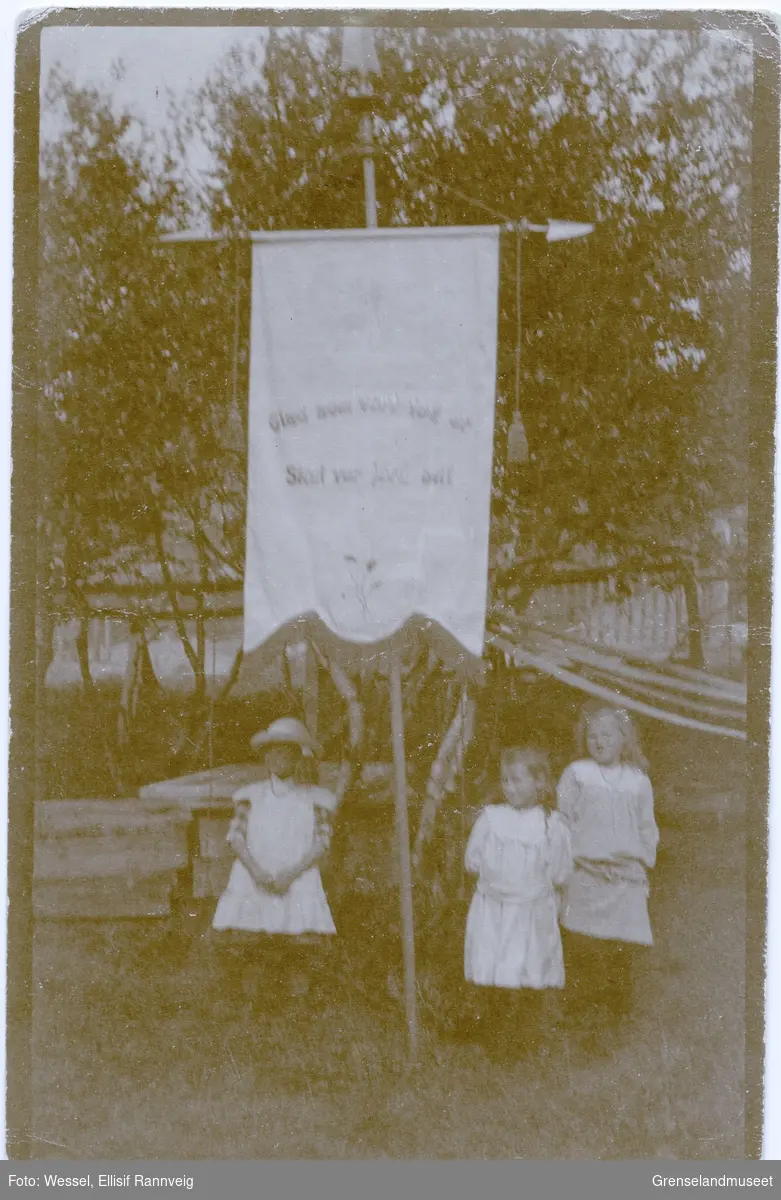 Barnas 1. mai i Kirkenes. Tre jenter står i en hage, foran en fane med inskripsjon: "Glad som vort tog er skal vor jord bli."