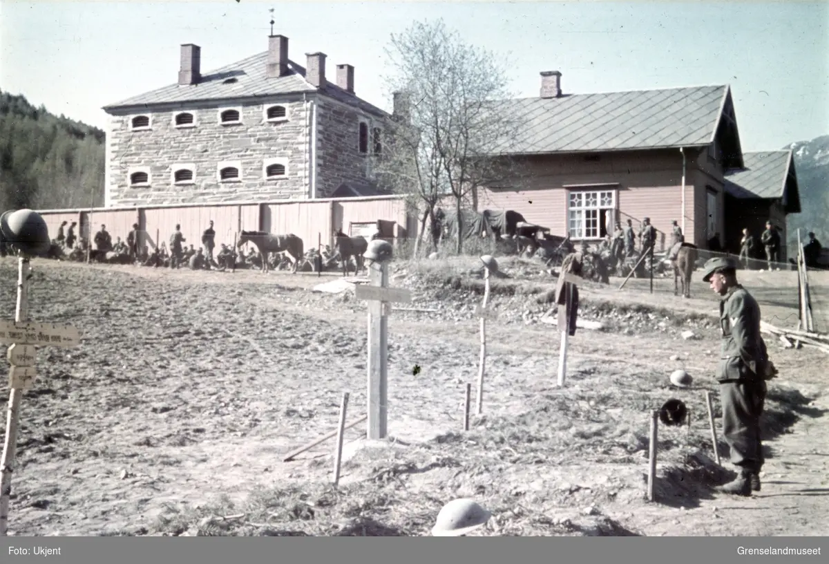 En tysk soldat står foran et jorde med flere graver. Tre av dem med tekst på. Hjelmene på korsene er av ulik type. I bakgrunnen ligger to bygninger, en i tre og en i stein. Rundt dem oppholder deg seg flere soldater, med hester og annet utstyr.