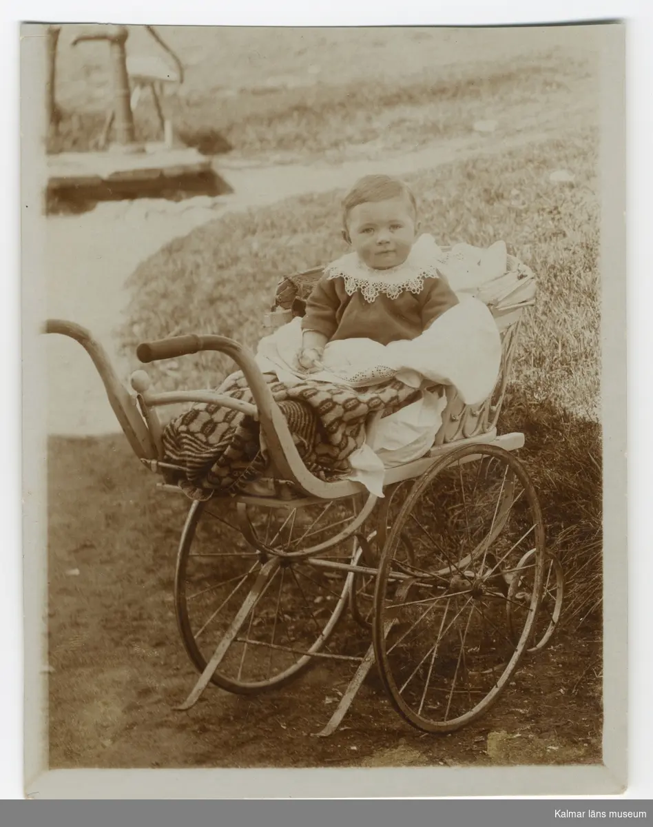 KLM 45133:3. Fotografi. Svartvitt. Motiv: Ett litet barn i en barnvagn/sittvagn med sufflett, i bakgrunden en handdriven vattenpump. Adresseringslinjer på baksidan. Odaterat. Sannolikt tidigt 1900-tal.