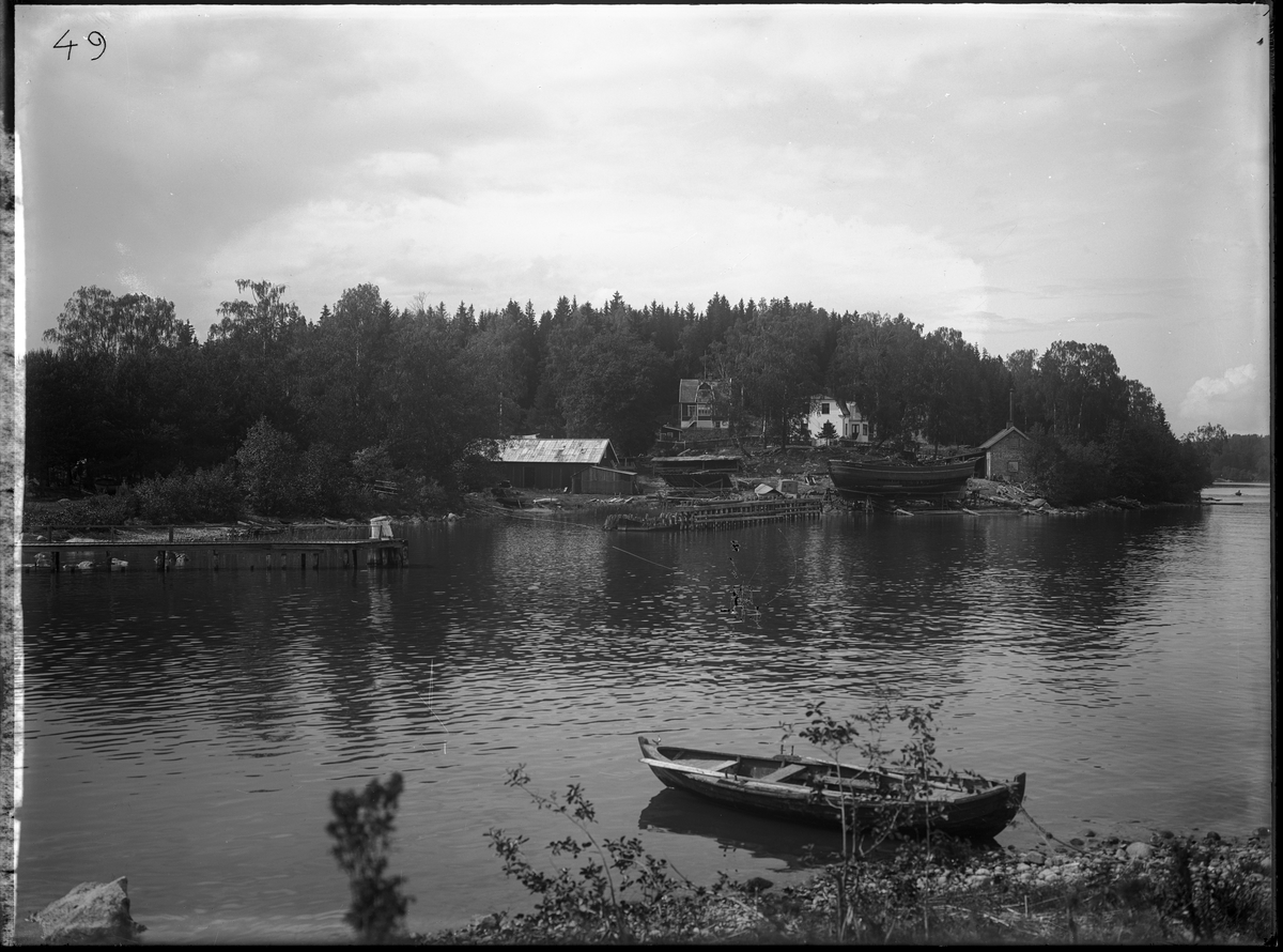 Fotografi taget i samband med bygget av Södertälje kanalverk. I bild syns ett par hus, en brygga, en eka, några småbåtar och skrovet till en större båt ståendes på land.