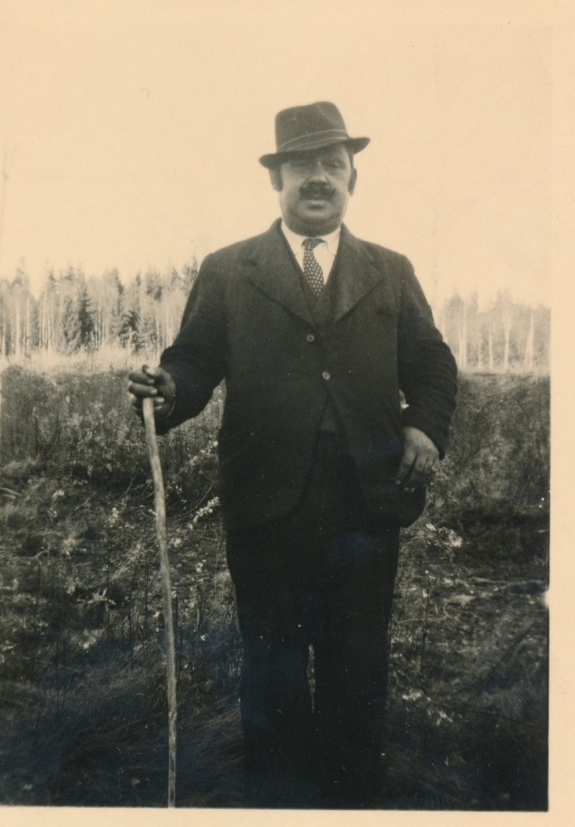 En man med hatt och käpp avporträtterad i ett skogslandskap.