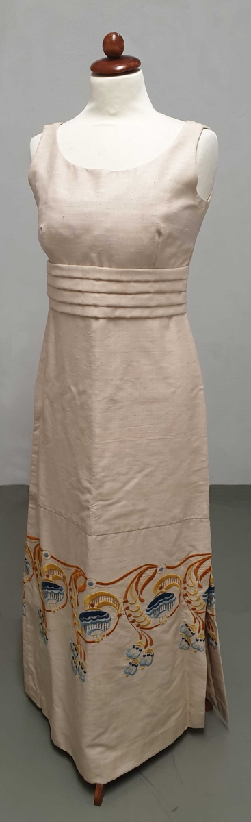 Beige ermeløs kjole av silke, med brodering i blått, brunt og gull langs skjørtekanten og med folder rundt rundt livet.