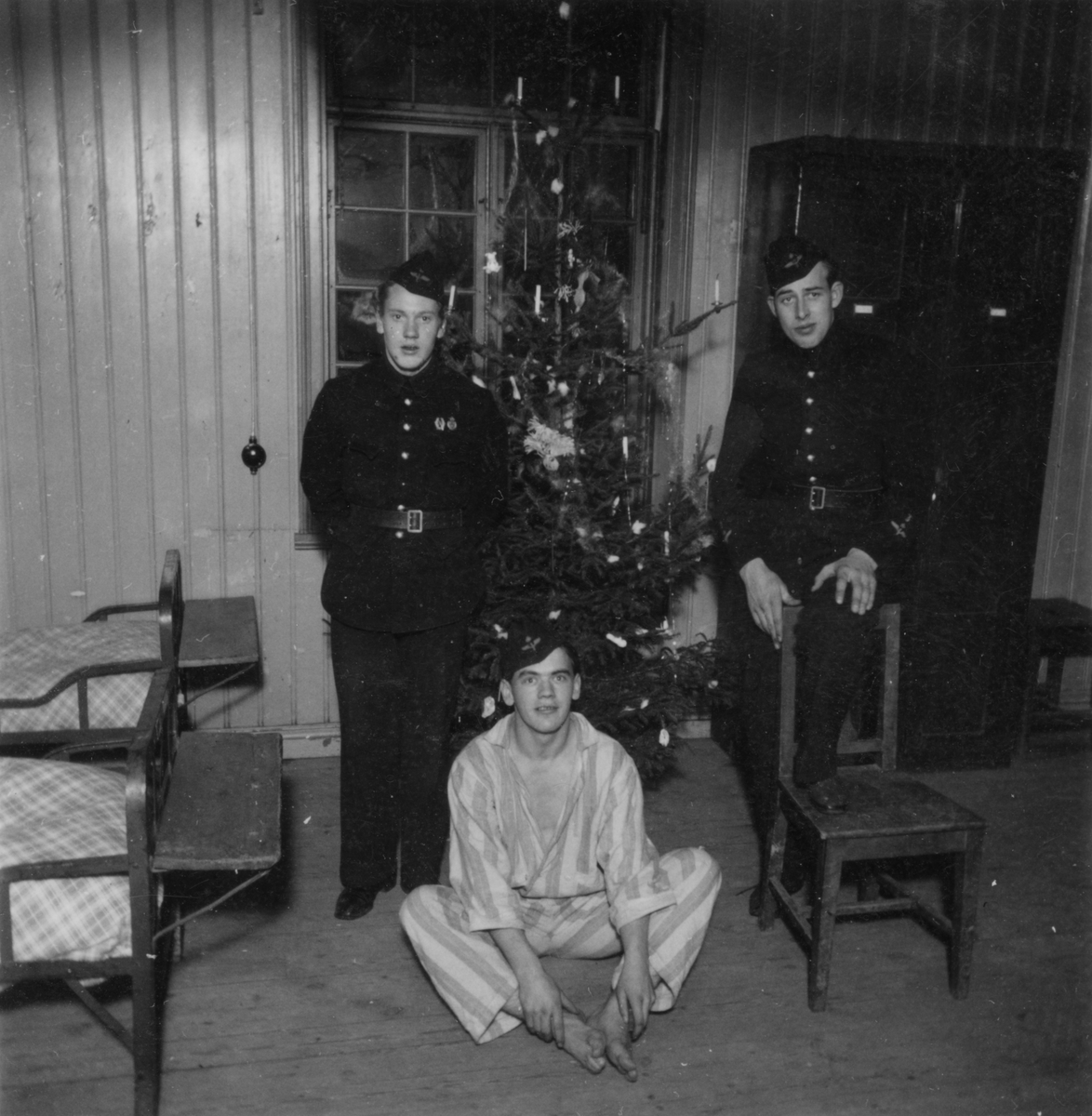 Grupporträtt av tre värnpliktiga män på logemente på F 3 Malmen, vid juletid. Två är klädda i flygvapenuniform och står vid en julgran. En är klädd i pyjamas och flygvapenmössa och sitter på golvet.