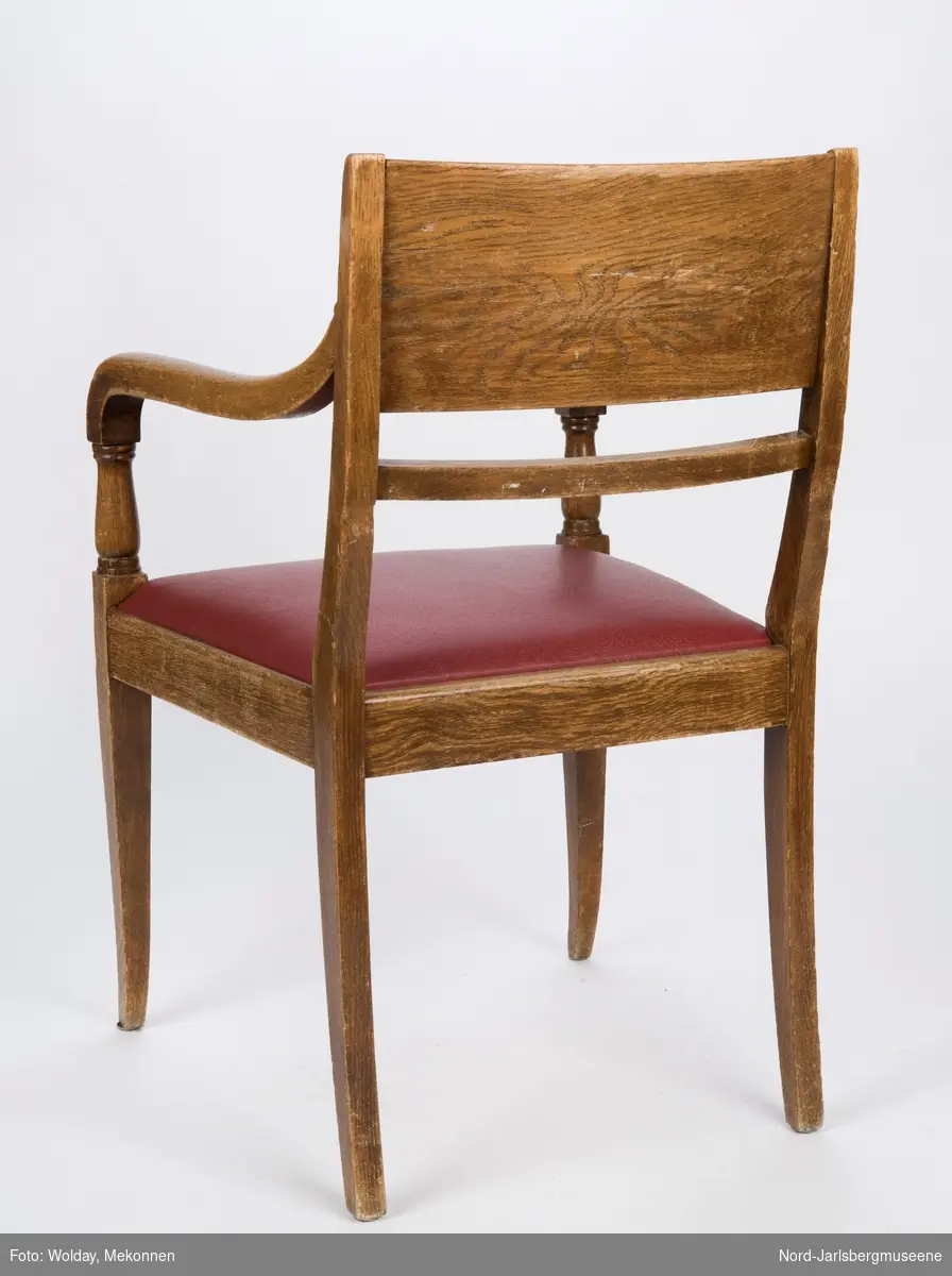 Stol i heltre med stoppet sete med rødt trekk. Stolen har forseggjorte armlener som avsluttes i en myk bue, med dreide detaljer. Ryggstø har en bred plate øverst og en tynnere list under den.