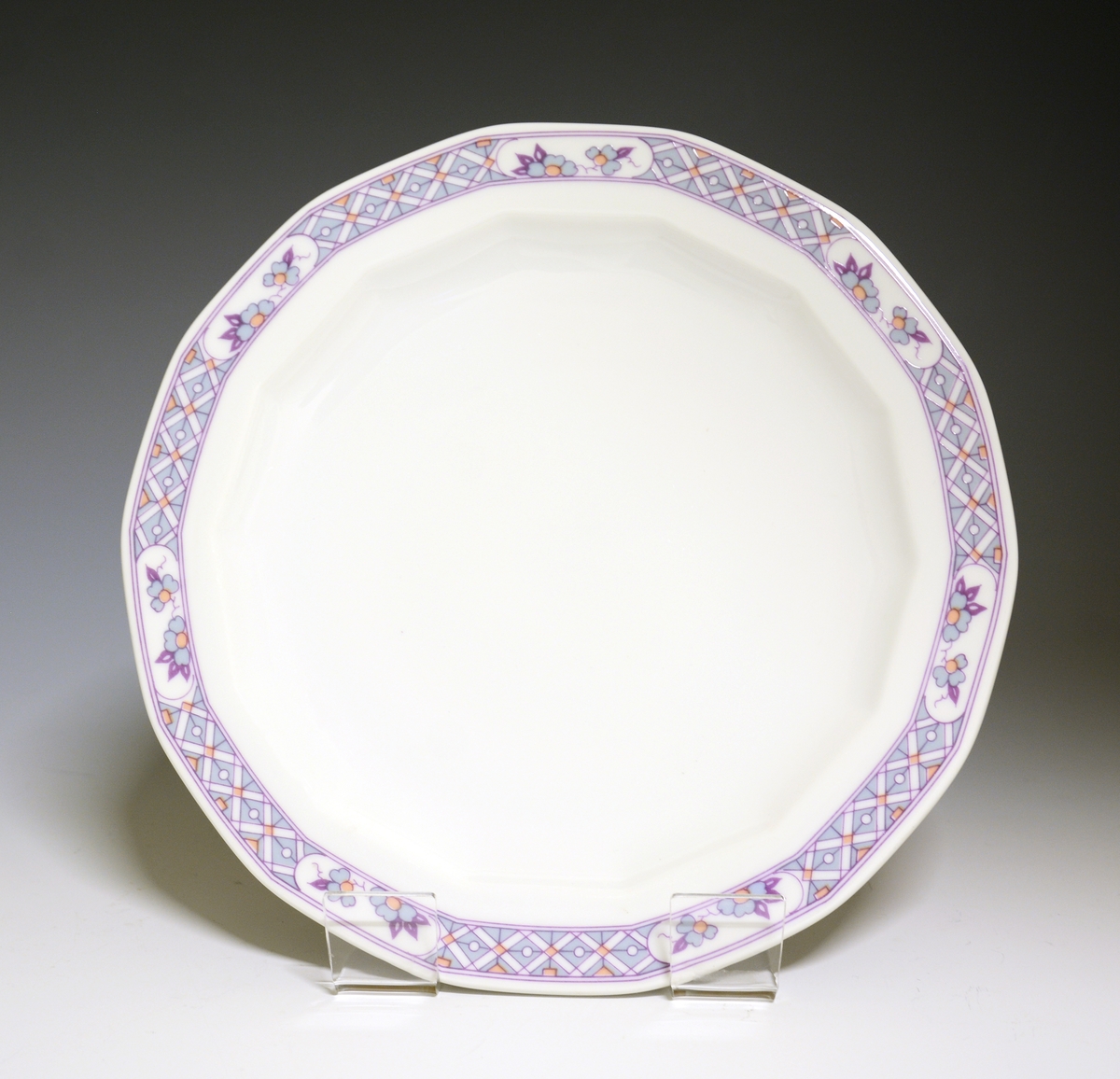 Mangekantet tallerken av porselen med hvit glasur. Dekorert med en bord ytterst på fanen med blomster og geometriske former. 
Modell: Octavia, i produksjon fra 1977
Dekor: Innkjøpt dekor, ikke av Grete Rønning.