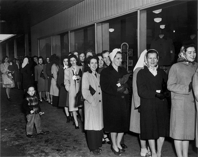 Bildet viser kvinner som står i kø og venter på å kjøpe nylonstrømper. Bildet er i svart/hvitt og alle kvinnene har på seg kåpe.