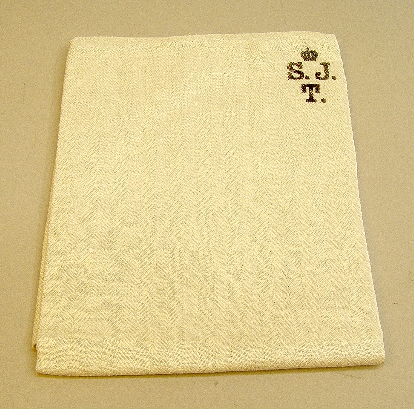 Rektangulär handduk av naturfärgat oblekt linne, troligtvis av äldre typ av handdukens utseende att döma. I två av hörnen är tryckt: S.J och bokstaven T nedanför. (= Statens Järnvägstrafik). På båda kortsidor hankar av vita bomullsband.