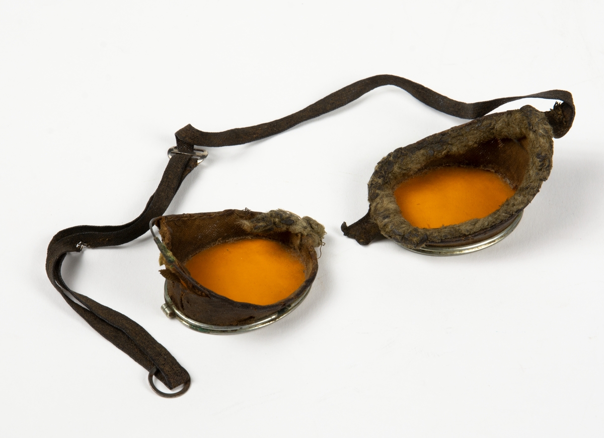 Flygglasögon med gult glas, sidskydd i tyg samt en rem att fästa glasögonen med på huvudet. Runt glasögonens innerkant finns en pälsliknande tyg.
Glasögonen har gått av i mitten och är i två delar.