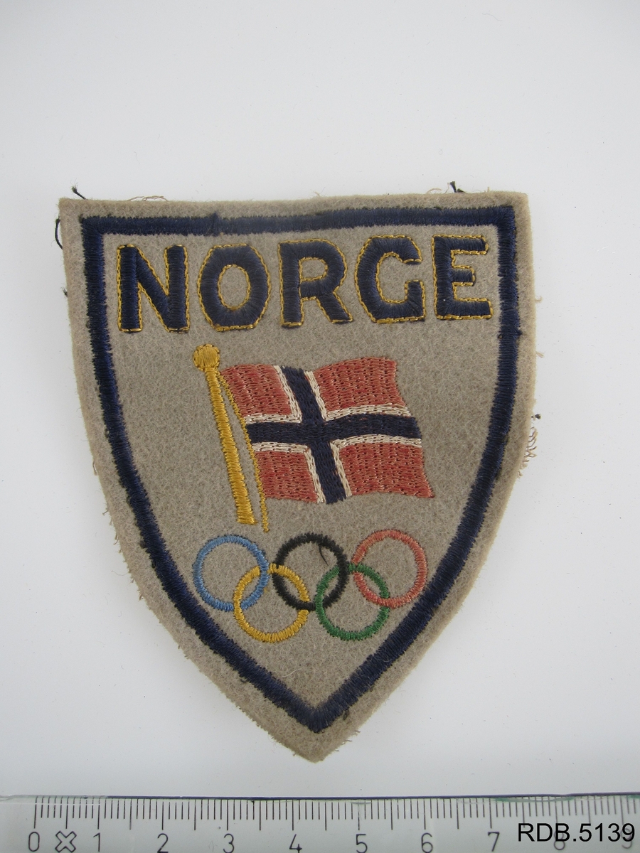 Trekantet jakkemerke i stoff. Grå bunnfarge. To forskjellige motiv; det norske flagg og OL-ringene. Norge i store bokstaver.