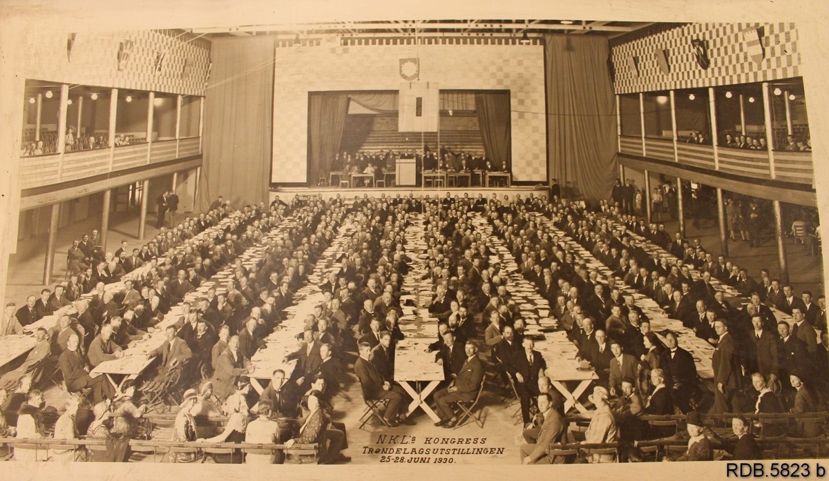 En stor forsamling menn og noen kvinner, deltakere på NKL's kongress under Trøndelagsutstillingen i 25.-28. juni 1930. Mennene sitter med papirer foran seg langs 7 lange bord . Det er også en scene fremst i salen og balkonger på hver side hvor det også sitter mennesker.