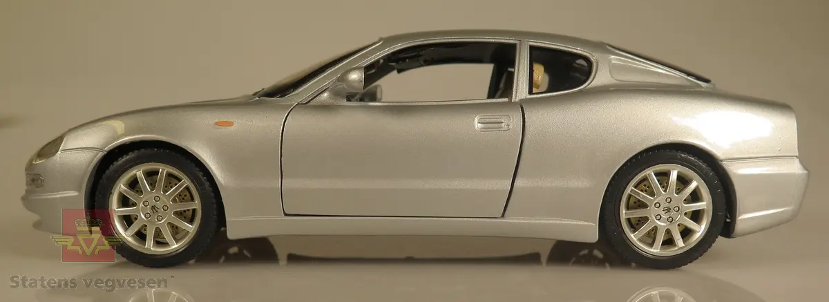 Primært grå modellbil laget av metall og er høyt detaljert. Den er utstyrt med funksjonellt ratt i tillegg til dører og bagasjelokk som kan åpnes og lukkes. Skala: 1:18