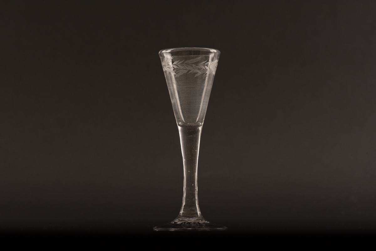 Brännvinsglas på rund fot av ofärgat glas. Kupan är koniskt formad och dekorerad med en ingraverad växtranka under mynningen.