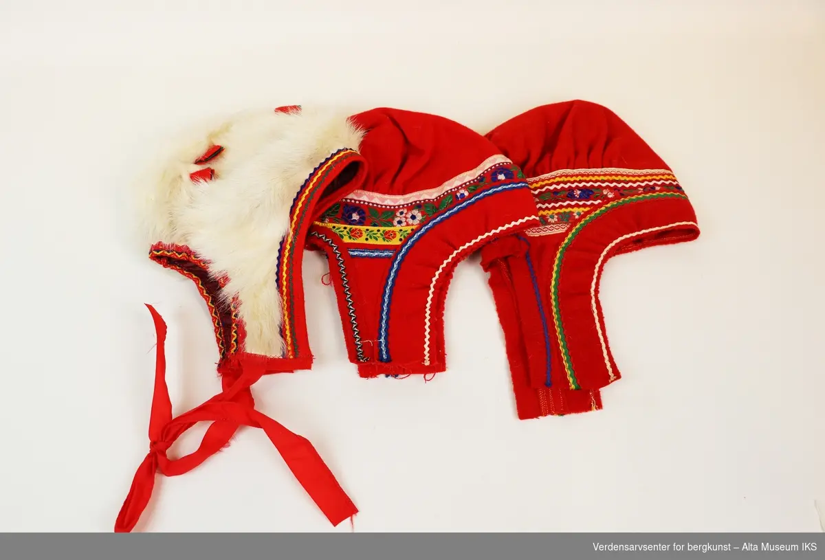 Samling dukkeklær med kofte fra Kautokeino, ett hvitt og ett mønstret sjal, rødt belte med dekorasjonsbånd, samt to røde sommerluer og ei vinterlue i hvit pels.