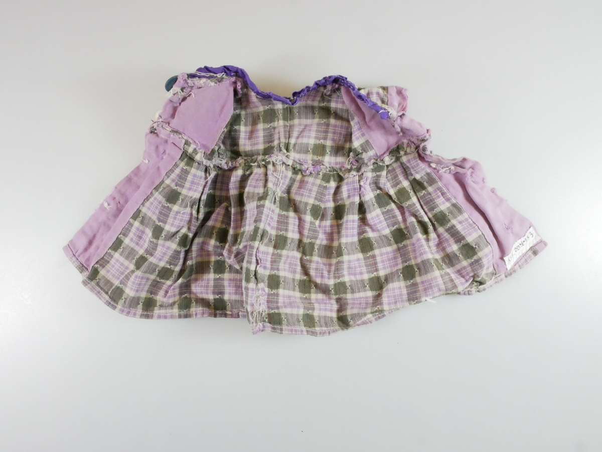Kortermet liten kjole i lilla og grønnrutete mønster, antageligvis brukt som dukkeklær. Tre hvite og en blå knapp på forstykker. 