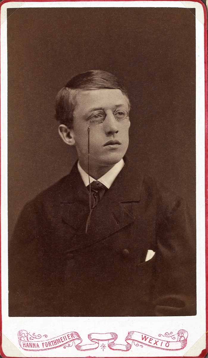Porträttfoto av en ung man med pincené, klädd i mörk kavajkostym med slips.
Bröstbild, halvprofil. Ateljéfoto.