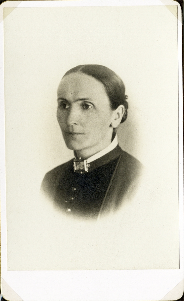 Porträttfoto av en kvinna i mörk klänning med vit ståkrage. Vid kragen syns en rektangulär brosch. 
Bröstbild, halvprofil. Ateljéfoto.