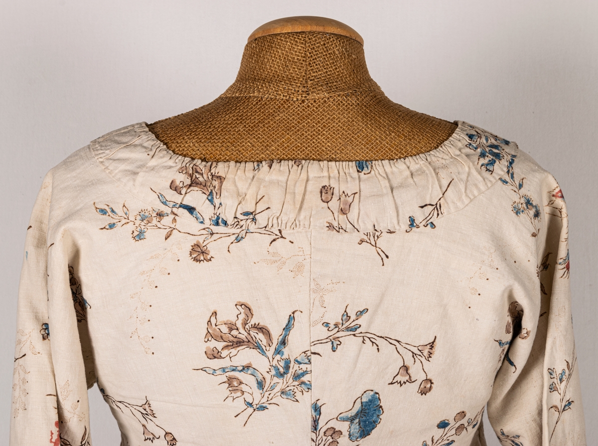 Tvådelad klänning, kjol och jacka, i naturfärgat linne med handtyckt blommönster i rött, brunt och blått. Klänningen är från 1700-talet.
a) jacka, urringad, knäppning fram, långa ärmar. Fodrad med naturfärgat linne. Lite svängd nedtill.
b) kjol, hellång, ofodrad, lagda vecka och smal linning.