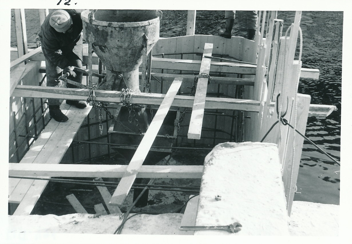 Mann i arbeid med betongstøping i forskalingskasse av tre og armeringsjern. I forbindelse at tverrgående mur ved Kistefos Træsliberi i Randselva repareres.