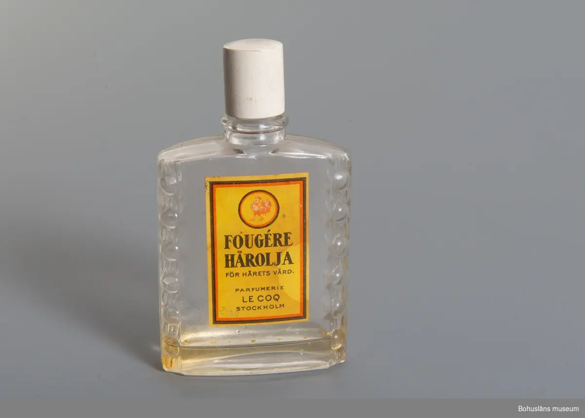 Oval flaska med bakelitskruvlock. 
Etikett i gult, rött,svart: "Fougére  hårolja för hårets vård Parfumerie Le Coq Stockholm."