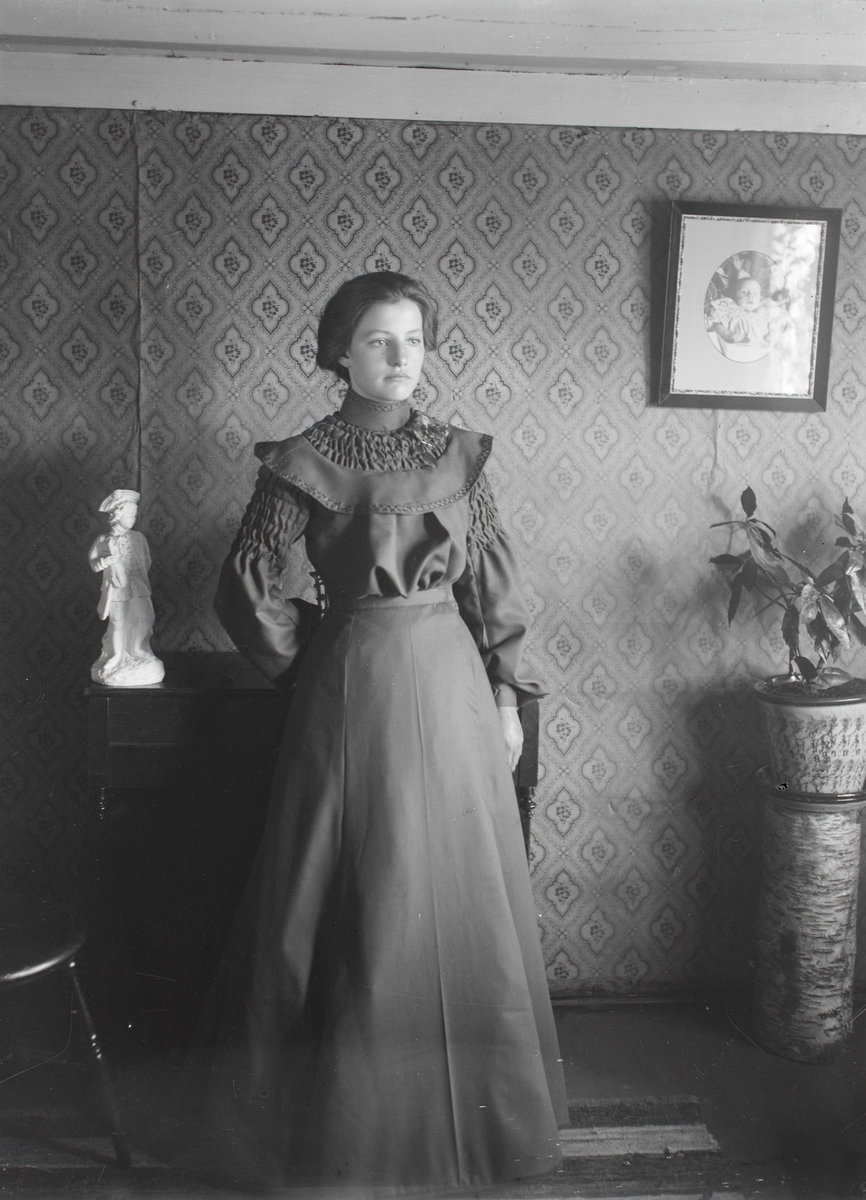 Portrett av ei kvinne i kjole som står i ei stue.