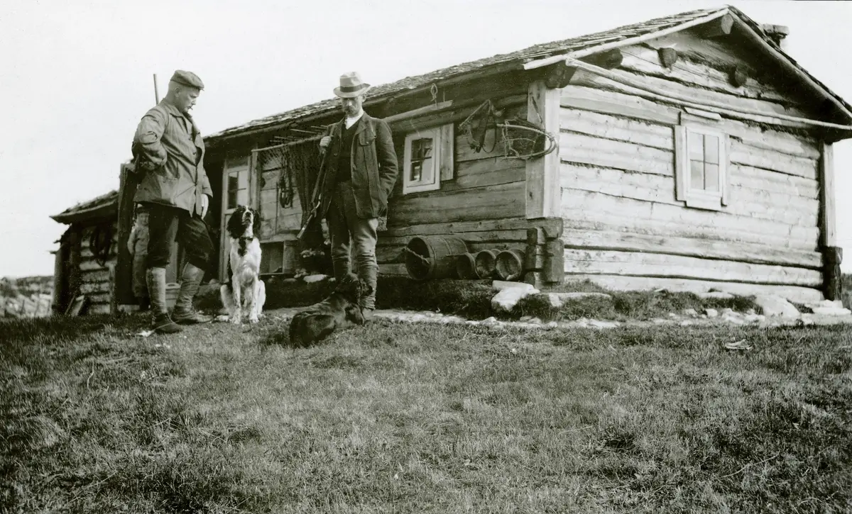 To jegere med fuglehund fotografert utenfor ei sæterstue i Lillerøåsen i Engerdal, som var tilholdssted for et jaktlag som leide terreng her av Trysil kommune. Jaktlaget besto av juristen Julius Thaulow Aubert, agent Louis Lühnenschloss, prokurist Heinrich Øhlckers, politisjef Johan Olaus Søhr, biblioteksjef Haakon Nyhuus og sakfører Sigurd Svenneby. Fotografiet ble tatt i 1910. Påfølgende år reiste kommunen ei ny jakthytte til dem. Leieavtalen hadde stått i tryslingen Haakon Nyhus’ navn. Etter at han var død tidoblet enka hans jaktavgiften til de øvrige jegerne, fra 60 til 600 kroner. Det ble i drøyeste laget for jegerne, men kjendisen Fridtjof Nansen hadde råd. Han betalte de 600 kronene. I løpet av to-tre uker skal polarhelten og jaktkameraten hans ha skutt 6-700 ryper i dette terrenget.